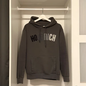 Mörkgrå hoodie från Hoodrich - Säljer en mörkgrå hoodie från Hoodrich med broderad logga i svart och grått framtill. Tröjan har huva med snörning, känguruficka och långa ärmar. Perfekt för en chill och street-inspirerad look. Pris kan diskuteras😊