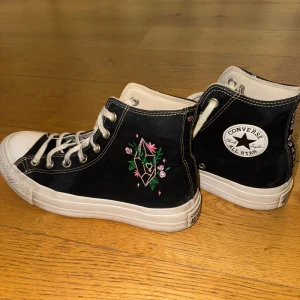 Converse All Star Rose Quartz High Tops - Svarta Converse All Star high tops med vita snören och platåsula, broderade blommor och geometriskt motiv på sidan samt texten 'Rose Quartz' broderad bak. Yttersulan är rosa. Skorna är i tyg och har klassisk rund tå och platå-sula. Använda ungefär en sommar 
