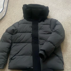 Svart Moncler Madeira puffer jacka - Säljer en svart pufferjacka från Moncler med stor vadderad huva och bred logotypremsa framtill. Jackan har en clean look, diskreta knappar och är perfekt för kalla dagar. Klassisk Moncler-stil med riktigt skön passform.