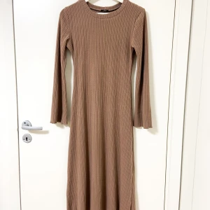 Ribbad beige långklänning från MiXMi - Supermysig ribbad långklänning i brun nyans från MiXMi. Klänningen har lång ärm, rund halsringning och medföljande bälte i samma material för att knyta i midjan. Perfekt för dig som gillar en enkel men stilren look.