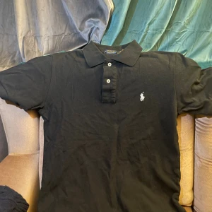 Svart pikétröja från Ralph Lauren - Svart pikétröja från Polo Ralph Lauren i modellen Custom Slim Fit. Klassisk krage, två knappar och den ikoniska broderade loggan på bröstet. Säljer den då jag har ingen behov av den så varför inte sälja den till ksk någon som gör!!!