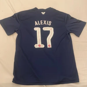 Arsenal Alexis 17 blå bortatröja - Snygg Arsenal bortatröja med Alexis 17 på ryggen. Tröjan är marinblå med färgblock i vitt, blått, turkost och brunt framtill. Klassisk v-ringning, kortärmad och tillverkad i lätt polyester. Arsenal-emblem och Fly Emirates-tryck på bröstet. Pris kan diskuteras 