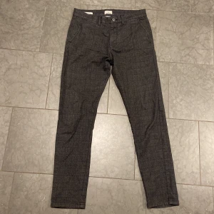 Grå rutiga kostymbyxor Jack & Jones - Slim fit kostymbyxor från Jack & Jones i mörkgrått med diskret rutmönster. Modellen heter Marco och har klassiska fickor fram och bak, bälteshällor och knappgylf. Perfekta för en clean och stilren look.
