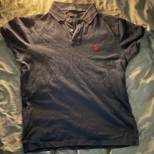 Mörkblå Polo Ralph Lauren piké - Snygg mörkblå pikétröja från Polo Ralph Lauren med klassisk krage och två knappar framtill. Den har det ikoniska röda broderade polo-spelaren på bröstet. Tillverkad i mjuk bomull som är skön mot huden och perfekt för en chill stil.