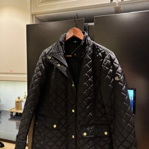 Svart quiltad jacka Lauren Ralph Lauren - Svart quiltad jacka från Lauren Ralph Lauren med guldiga RL-detaljer på knappar och fickor. Klassisk design med lång ärm, hög krage och två stora fickor framtill. Snygg och stilren jacka som passar perfekt till höst och vår.