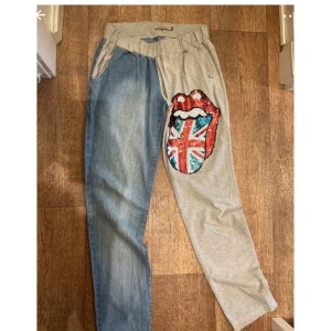 Unika tvåfärgade byxor med tryck - Snygga byxor med ena benet i ljusblå denim och andra i grå bomull. Framsidan har ett stort tryck av läppar med Union Jack-flagga i paljetter. Resår i midjan och fickor på sidorna. Perfekt för dig som vill sticka ut med en cool och annorlunda stil.