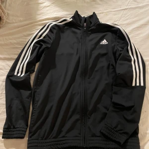 Svart Adidas track jacket med vita ränder - Svart track jacket från Adidas med klassiska vita ränder längs ärmarna och Adidas-logga på bröstet. Jackan har hel dragkedja framtill, hög krage och långa ärmar med resår vid ärmslut och nederkant. Perfekt för en sportig och avslappnad stil. XL i barnstorlek men har använts av en man som bär storlek S-M  och nassar kvinna i storlek M.
