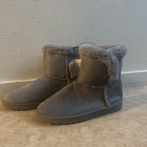 Grå fluffiga boots med foder - Mysiga grå boots med mjukt foder och mockaliknande utsida. Skorna har rund tå, platt sula och fluffig kant upptill. Perfekta för kalla dagar när du vill hålla fötterna varma och bekväma.