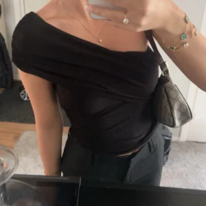 Svart oneshoulder topp i omlottstil - Säljer en svart oneshoulder topp med snygg omlott-effekt och draperade detaljer. Toppens tighta passform framhäver siluetten och den är tillverkad i ett mjukt, stretchigt material som sitter bekvämt. Perfekt för dig som gillar stilrena och trendiga plagg.