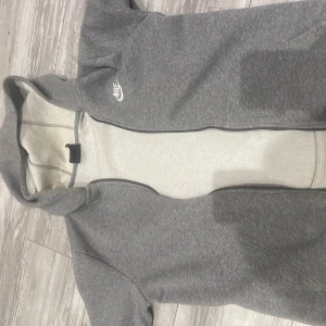 Grå hoodie från Nike i fleece  - Mysig grå hoodie från Nike i storlek XL med vit Nike-logga på bröstet. Tröjan har dragkedja, huva och är fodrad med mjuk fleece på insidan. Perfekt för kyliga dagar och chill vibes.