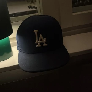 Blå LA-keps från New Era - Säljer en klassisk blå keps från New Era med LA-logga framtill i vitt. Kepsen har rak skärm och broderade detaljer på sidorna och bak. Perfekt för dig som gillar sportig stil och vill ha en ikonisk accessoar.