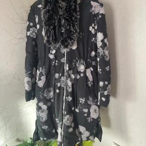 Blommig svart dunjacka med fuskpäls strl M - Snygg svart dunjacka med vita blommor och avtagbar fuskpäls runt kragen. Jackan har dragkedja framtill, praktiska fickor och huva. Perfekt för kalla dagar när du vill sticka ut lite extra. Rymlig i storlek. 38/M. Märke är Mohito.
