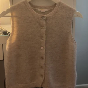 Beige stickad väst från Gina Tricot - Mysig beige stickad väst från Gina Tricot med rund hals och knappar framtill. Västen har en enkel och clean look med ribbade kanter och är perfekt att styla över en t-shirt eller skjorta. Passar dig som gillar en chill och klassisk vibe.aldrig använt!!så är i väldigt bra skick.