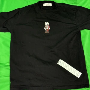 Offwhite svart T-shirt  - Offwhite Svart t-shirt med en liten figur på framsidan och en större tryckt figur på baksidan. Figuren på baksidan har en komplex design med olika element. Liknande modell som originalet!