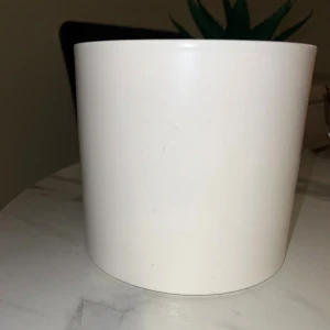 Kruka i bra skick  - Bilderna visar en vit, cylindrisk kruka i keramik, cirka 16 cm hög och 17 cm i diameter. Ytan är slät med några mindre repor och märken, men i gott skick. Perfekt för inomhusväxter eller dekoration. Enkel och stilren design som passar de flesta hem.
