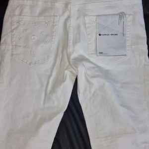 Vita jeans från Purple Brand, strl 38 - Säljer ett par fräscha vita jeans från Purple Brand i storlek 38. Byxorna har snygga broderade detaljer på bakfickorna och längs benen, klassisk femficksmodell och silverfärgade nitar. Perfekt för dig som vill sticka ut med stilrena detaljer.