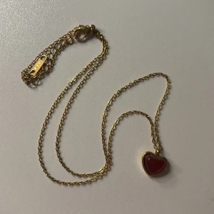 Guldhalsband med rött hjärthänge ❤️ - Supersött halsband i guld med ett rött hjärta som hänge. Kedjan är tunn och elegant med justerbar längd. Hjärtat har en glansig finish och är inramat i guldtonad metall, vilket ger en romantisk och trendig vibe. Perfekt accessoar för att lyfta vilken outfit som helst.