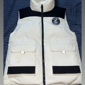 Vit och svart dunväst från Canada coouse - Snygg dunväst i vitt med svarta detaljer från Balabala. Västen har hög krage, avtagbar huva och två stora fickor framtill med svarta paneler. Dragkedja hela vägen och coolt märke på bröstet. Perfekt för lager-på-lager under kyliga dagar.