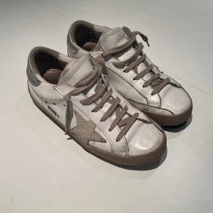 Vita golden goose’s skor  - Snygga vita sneakers från Golden Goose med grå mockadetaljer och en ikonisk stjärna på sidan. Skorna har grå snörning, beige sula och svart yttersula. Tillverkade i skinn och mocka för en cool och avslappnad look. Dom är lite förstöra skriv om ni vill ha bild är även öppen för byten inom stockholm.