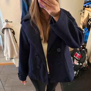 Mörkblå skepparkavaj med knappar - Säljer en klassisk mörkblå skepparkavaj med dubbelknäppning och bred krage. Jackan har stora knappar framtill och på ärmsluten, samt två fickor. Perfekt för dig som vill ha en stilren och tidlös look.