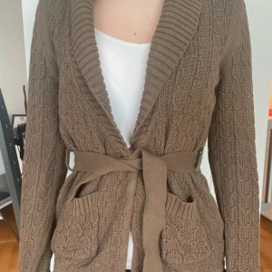 Beige stickad kofta med bälte - Mysig beige stickad kofta med snyggt strukturmönster, bred ribbad krage och knytskärp i midjan. 