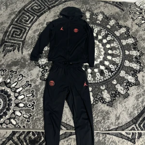 Svart PSG x Jordan träningsset - Svart hoodie från Jordan x PSG med röd Jumpman-logga och PSG-märke på bröstet. Tröjan har huva, hel dragkedja och långa ärmar. Perfekt för dig som gillar sportig och clean stil.