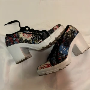 Blommiga sneakers med klack - Unika sneakers med chunky vit klack och blommigt mönster i blått, rött och vitt. Skorna har snörning och en plattformsula som ger extra höjd. Perfekta för dig som vill sticka ut med färg och cool design.
