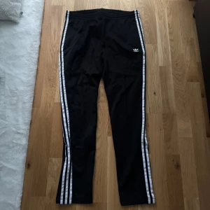 Svarta Adidas byxor med vita ränder - Svarta Adidas byxor med klassiska vita ränder längs sidorna och Adidas-logga på ena höften. Tillverkade i Primeblue-material, vilket innebär återvunnen polyester. Byxorna har elastisk midja och sportig look, perfekta för en avslappnad stil. Dragkedjor på sidofickorna. 