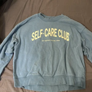 Blå sweatshirt  - Blå sweatshirt med trycket 'SELF-CARE CLUB' i gult framtill. Tröjan har rund hals, långa ärmar och ribbade muddar vid ärmslut och nederkant. Perfekt för chill dagar eller när du vill ha en avslappnad look.