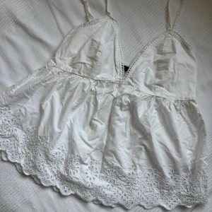 Vit broderad linnetopp från BikBok - Supersöt vit linnetopp från BikBok med smala axelband, v-ringning och broderade blommor längs den vågiga nederkanten. Toppen har smock i ryggen för extra komfort och en luftig passform, perfekt för varma dagar.
