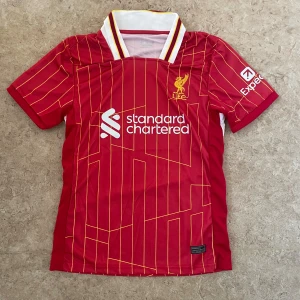 Liverpool FC röd fotbollströja - Snygg röd Liverpool FC fotbollströja med gula och vita detaljer, samt tunna gula ränder över hela tröjan. Kortärmad modell med vit krage och klubbmärke på bröstet. Tillverkad i lätt och ventilerande material, perfekt för match eller träning.