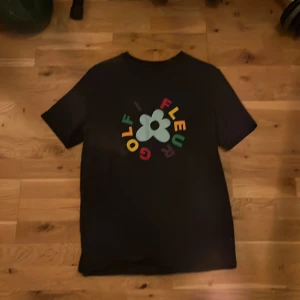 Golf Le Fleur svart t-shirt med tryck - Svart t-shirt från Golf Le Fleur med färgglatt blomtryck och text på bröstet. Klassisk passform och rund hals. Tillverkad i 100% bomull för skön känsla. Perfekt för dig som gillar streetwear och vill sticka ut med en ikonisk design.