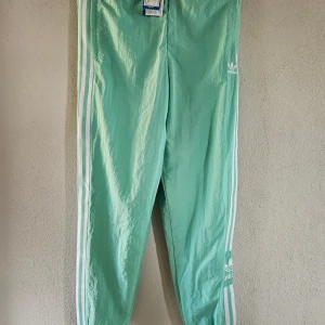 Mintgröna Adidas byxor med vita ränder - Snygga mintgröna Adidas byxor med klassiska vita ränder längs sidorna och broderad logga på låret. Modellen heter Lock Up TP och har elastisk midja. Perfekta för en sportig och avslappnad stil.