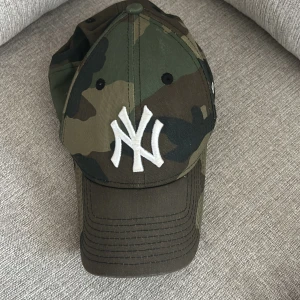 NY Yankees keps camo New Era 9FORTY - Säljer en New Era 9FORTY keps med New York Yankees-logga broderad framtill. Kepsen har ett snyggt camouflage-mönster i gröna och bruna toner och är tillverkad i 100% bomull. Justerbar passform baktill. Perfekt för dig som gillar streetwear och sportig stil.
