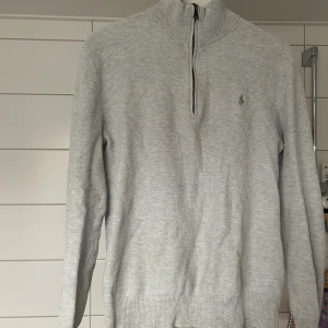Ljusgrå half zip tröja Polo Ralph Lauren - Ljusgrå stickad tröja från Polo Ralph Lauren i mjuk pima bomull. Klassisk modell med half zip, hög krage och diskret broderad logga på bröstet. Långärmad och ribbade muddar för en clean look. Perfekt för lager-på-lager under höst och vinter.har använts under lång tid men är i okej skick. Tidigare hål i armhålan men är lagat nu. 