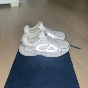 Vita Dior sneakers med CD-detalj - Snygga vita sneakers från Dior med reflekterande CD-logga på sidan och bak. Skorna har en chunky sula, snörning och är tillverkade i en mix av mesh och skinn för en lyxig känsla. Perfekt för dig som gillar exklusiva märken och stilren design.