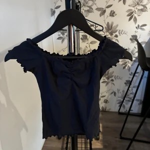 Svart topp - Svart ribbad topp från Hollister med söt rynkdetalj framtill och volangkant vid halsringning, ärmslut och nederkant. Modellen är figurnära och har korta ärmar med offshoulder-stil. Perfekt för dig som gillar en enkel men ändå trendig look.