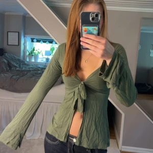 Olivgrön omlottopp med knyt - Olivgrön långärmad topp i mjuk bomull med snygg omlott och knyt framtill. Toppen har v-ringning och vida ärmar som ger en avslappnad vibe. Perfekt att styla med jeans eller kjol för en trendig look. Tården har gått upp på två ställen men går lätt att sy, de två sista bilderna. XS/S