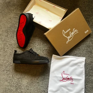 Christian Louboutin svarta sneakers med nitar - Säljer ett par svarta sneakers från Christian Louboutin med ikonisk röd sula och svarta nitar på tån. Skorna är i mocka med snörning och har en clean, edgy look som sticker ut. Kommer med originalkartong och dustbag.