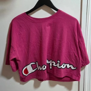 Champion Croppad T-shirt / Crop Top i Stark Fuchsia/Rosa Storlek M - Säljer en trendig och snygg Champion T-shirt  med croppad passform (Crop Top). T-shirten är i en härlig, pigg färg, bäst beskriven som stark rosa eller fuchsia, och har Champions klassiska stora logotyp tryckt över bröstet. Perfekt för gymmet, sportiga outfits eller till höga byxor och shorts.  Märke: Champion Storlek: M (Medium) Färg: Fuchsia / Stark Rosa Modell: Croppad (Loose fit, kort) Skick: Mycket fint skick.