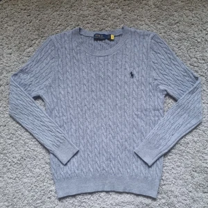 Grå kabelstickad tröja Polo Ralph Lauren - Kabelstickad grå tröja från Polo Ralph Lauren med rund halsringning och broderad logga på bröstet. Tröjan har långa ärmar och ribbade muddar. Perfekt för lager-på-lager och snyggt nu på vintern. Tröjan är orginellt L (Large) men har krympt i tvätten så skulle säga att den är S (Small) numera. Använt 1 gång. Nypris: 2395 kr. Mitt pris: 800. Accepterar bud.
