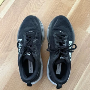 Svarta Hoka Bondi 8 sneakers - Säljer ett par svarta Hoka Bondi 8 sneakers med vit och grå tjock sula. Skorna har ventilerande mesh-ovandel, snörning och Hoka-logga på sidan. Strl 39 1/3. Använd endast 2 ggr