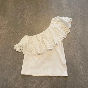 Vit offshoulder topp med volang - Säljer en söt vit offshoulder topp med bred volang och broderade blommor längs kanten. Toppen har en axel bar och är tillverkad i mjuk bomull. Perfekt för dig som gillar romantisk och feminin stil. Den är i storlek 152 men passar mig som vanligtvis bär xs/34.