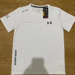 Vit Under Armour t-shirt (helt ny+gratis frakt) - Vit t-shirt från Under Armour med HeatGear-teknologi, perfekt för träning. Kortärmad modell med svart logga på bröstet och tryckt 'UNDER ARMOUR' längs sidan. Tillverkad i lätt och snabbtorkande polyester för maximal komfort.