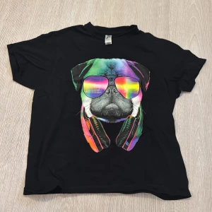 Svart t-shirt med färgglad hund från SOL'S - Cool svart t-shirt från SOL'S med en stor färgglad print av en mops med spegelglasögon och hörlurar runt halsen. T-shirten är kortärmad och i mjuk bomull, perfekt för dig som gillar unika och roliga motiv.