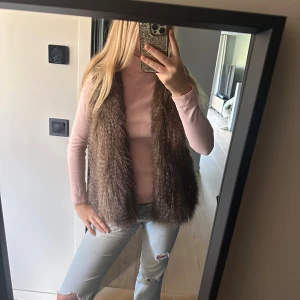 Brun fluffig pälsväst - Säljer en brun fluffig pälsväst med mjukt foder på insidan. Västen har en loose passform och är perfekt för att ge din outfit en cool och trendig touch. Färgen är naturligt brun med lite skiftningar och materialet är pälsimitation.