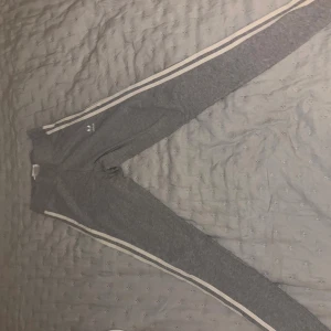 Grå Adidas mjukisbyxor med vita ränder - Grå mjukisbyxor från Adidas med klassiska vita ränder längs benen och broderad logga på höften. Byxorna har en slim passform och är tillverkade i mjuk bomullsmix. Perfekta för en sportig och avslappnad look.