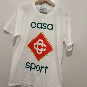 Vit Casablanca Casa Sport t-shirt M - Vit t-shirt från Casablanca med tryck 'casa sport' och en stor röd och gul grafisk logga på bröstet. Klassisk passform med korta ärmar och rund hals. Tillverkad i 100% bomull, perfekt för en sportig och clean look.