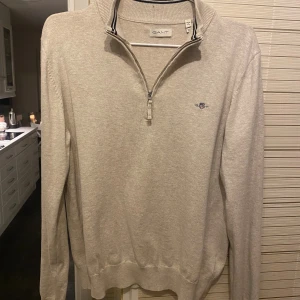 Beige half zip tröja från GANT - Snygg beige tröja från GANT med half zip och broderad logga på bröstet. Tröjan har lång ärm, ribbade muddar och en klassisk krage med dragkedja. Perfekt för lager-på-lager och stilren look. Storleken är 176cm. Har använt tröjan 1-3 gånger.