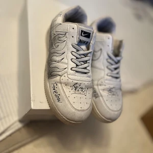 Nike Air Force 1 custom vit med svart doodle - Unika Nike Air Force 1 sneakers i vitt skinn med handritade svarta doodles och textdetaljer över hela skon.(Har använt de några gånger och det märks endast om man kollar under skorna)olKlassisk låg modell med snörning och rund tå. Perfekt för dig som vill sticka ut med en personlig touch på dina kicks.